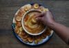 Recipes: How to Make Gandules Hummus A white bowl filled with Gandules Hummus garnished with achiote oil and sazón, surrounded by a ring of crispy homemade green plantain chips (platanutres) on a wooden board
