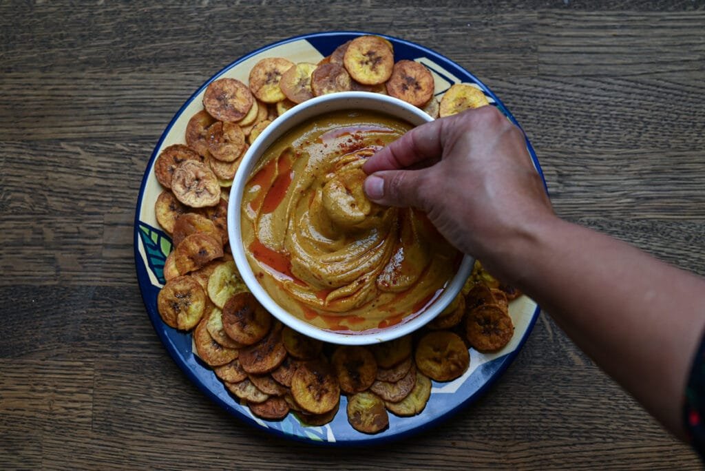A white bowl filled with Gandules Hummus garnished with achiote oil and sazón, surrounded by a ring of crispy homemade green plantain chips (platanutres) on a wooden board