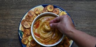Recipes: How to Make Gandules Hummus A white bowl filled with Gandules Hummus garnished with achiote oil and sazón, surrounded by a ring of crispy homemade green plantain chips (platanutres) on a wooden board