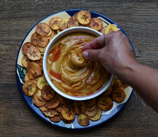 Recipes: How to Make Gandules Hummus A white bowl filled with Gandules Hummus garnished with achiote oil and sazón, surrounded by a ring of crispy homemade green plantain chips (platanutres) on a wooden board