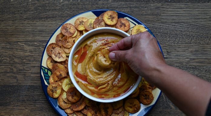 Recipes: How to Make Gandules Hummus A white bowl filled with Gandules Hummus garnished with achiote oil and sazón, surrounded by a ring of crispy homemade green plantain chips (platanutres) on a wooden board